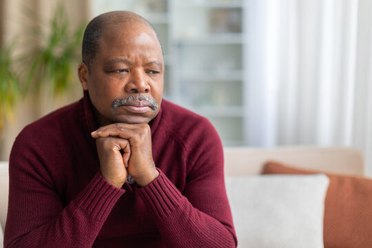 Thoughtful mature Black man sitting at home with his chin resting on clasped hands, looking away with a serious, concerned expression in a quiet modern living room interior