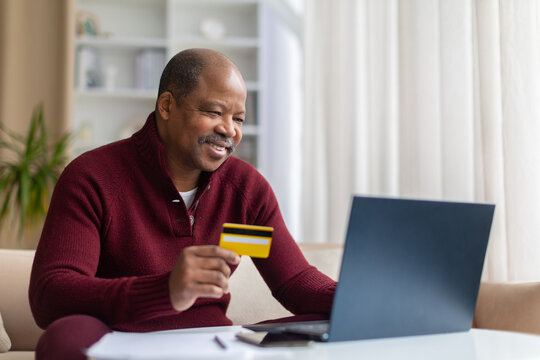 Smiling mature Black man using laptop and holding credit card at home, making an online purchase, payment or banking transaction from a comfortable living room