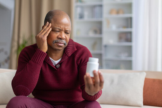 Mature Black man sitting on a sofa at home, holding a medicine bottle while touching his temple, illustrating headache, illness, medication use and home healthcare