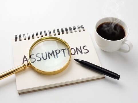 A Spiral Notepad Displaying The Word Assumptions Written In Black Marker Viewed Through A Brass Magnifying Glass Alongside A Black Pen And A Steaming Cup Of Coffee On A White Background