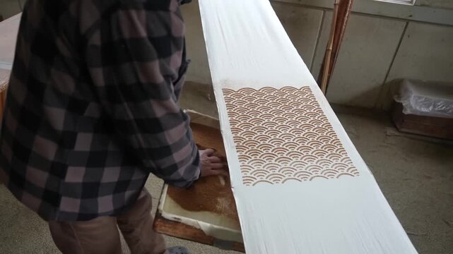 Dyer scatters fine sawdust from a shallow tray over freshly applied resist paste running the length of a long white cloth
