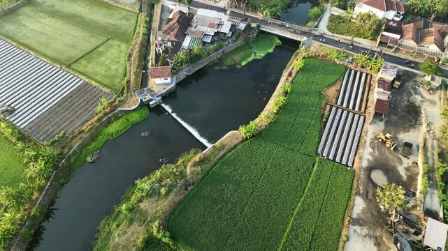 Static aerial drone video of a river with a small dam flowing through a rural village. Surrounding landscape features green agricultural fields, houses, and a road bridge under morning sunlight.