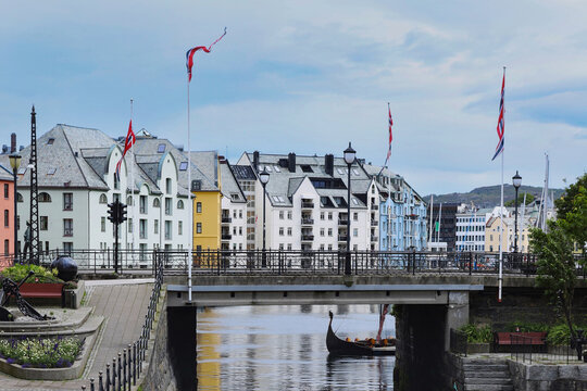 Scenic view of hellebroa bridge over the brosundet canal with norwegian flags. Traditional art nouveau buildings and traditional wooden viking boat on a cloudy day in alesund city.