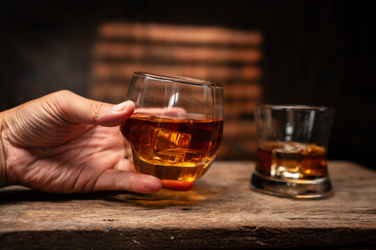 Hand Holding Glass of Whiskey with Ice on Wooden Table in Bar