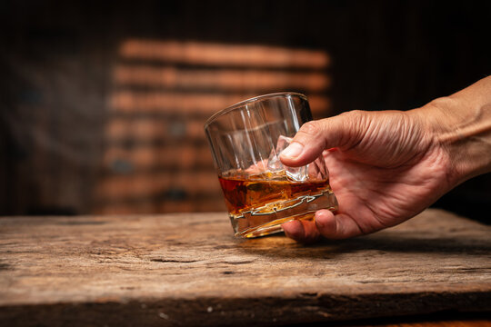 Hand Holding Glass of Whiskey with Ice Cubes on Wooden Table