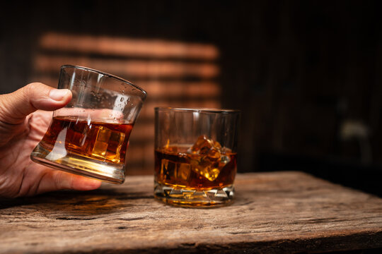 Hand Holding Glass of Whiskey with Ice on Wooden Table in Bar