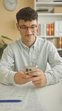 Young hispanic man wearing glasses holding smartphone, smiling and fist pumping over a highlighted spreadsheet at home office desk; happiness success.