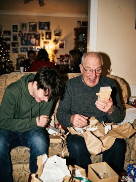Grandfather laughs with delight while opening a wrapped Christmas gift as his grandson giggles beside him at home.