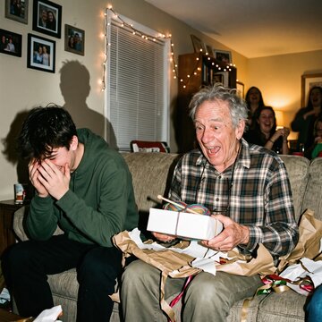 Grandfather laughs with delight while opening a wrapped Christmas gift as his grandson giggles beside him at home.