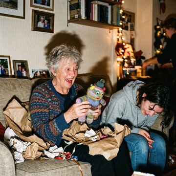 Grandmother laughs joyfully while unwrapping a knitted toy as her embarrassed granddaughter covers her face beside her.
