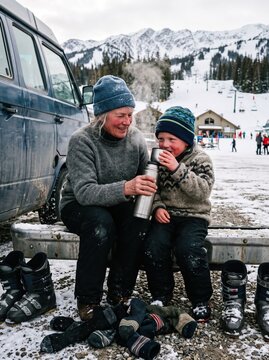 Grandmother pours a warm drink from a thermos for his laughing grandson beside a van at a snowy ski resort.