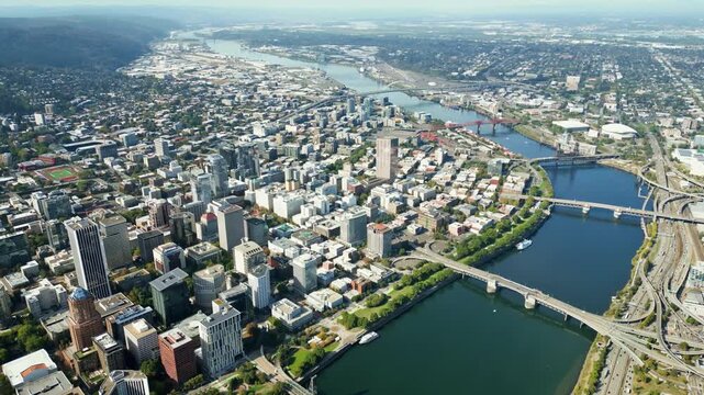 Aerial view of downtown Portland cityscape with Willamette River flowing through city, showcasing urban infrastructure and surrounding natural landscape on sunny day.