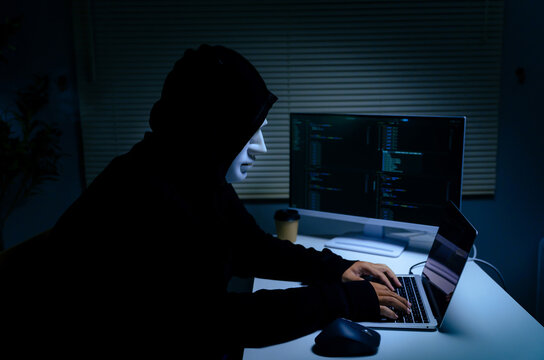Hacker identity theft concept wearing anonymity mask while using laptop for cybersecurity breach and data hacking