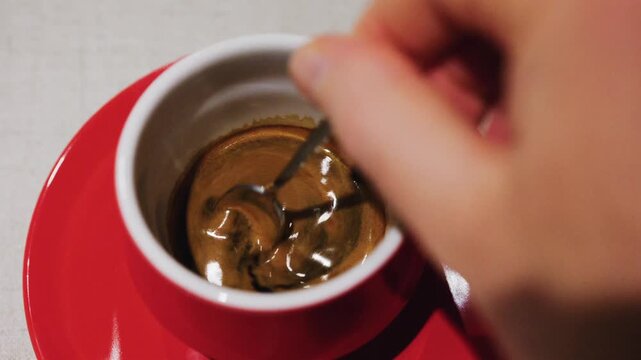 A person stirs a rich, dark espresso with a small spoon in a red cup, creating captivating swirling patterns in the creamy foam.