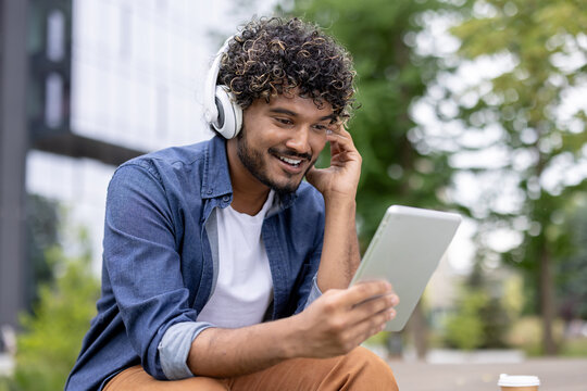 Young man with curly hair wearing headphones, smiling while looking at a tablet screen outdoors, sitting on a bench with a takeaway coffee, connecting with technology for leisure and communication