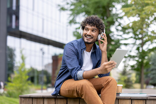 Young man with curly hair enjoying music through headphones, holding a digital tablet, and smiling while relaxing on a wooden bench in an urban park