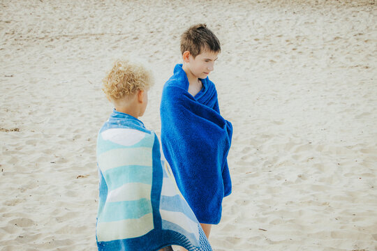 Two boys, wrapped in blue beach towels, stand together on the light sand after swimming.