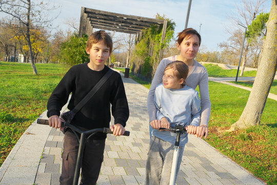 Mother with her young daughter and son riding kick scooters together along a paved path in a sunny park. Happy active family outdoor leisure