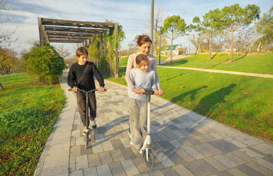 Mother with her young daughter and son riding kick scooters together along a paved path in a sunny park. Happy active family outdoor leisure