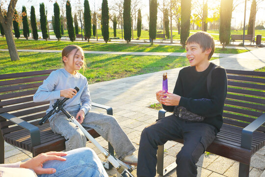 Laughing brother and sister holding a chocolate bar and a kick scooter while sitting on wooden benches in a park. Cheerful authentic children emotions