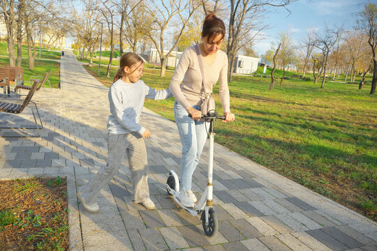 Smiling girl pushing her mother riding a white kick scooter along a paved path in a sunny park. Happy authentic family active