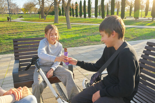 Smiling girl sharing a chocolate bar with her brother while sitting on benches in an autumn park. Authentic childhood sharing