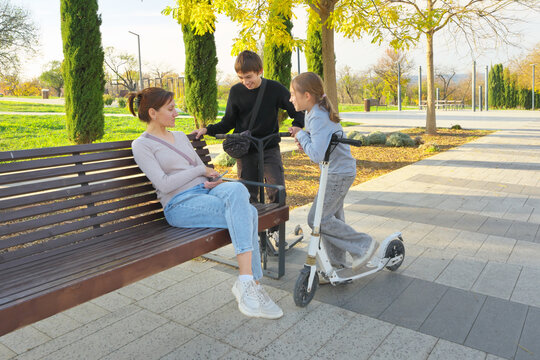 Mother with son and daughter laughing together near a wooden bench with kick scooters in an autumn park. Cheerful authentic family joy scene