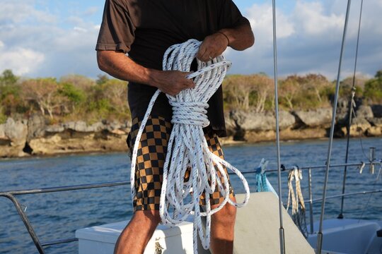Sailor coiling a mooring rope on a sailing yacht deck. Close-up of a man coiling a white nylon rope in his hands on the deck of a sailing yacht, with a rocky coastline and blue sea in the background. 