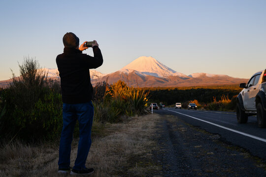 Tourist standing by the busy road and taking smartphone photos of Mt Ngauruhoe at sunset. Tongariro National Park. New Zealand.