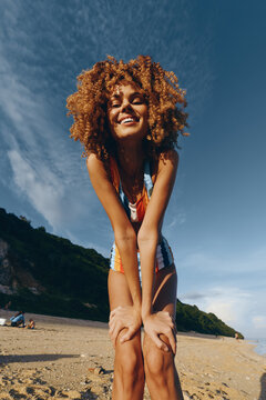 Young woman with curly hair in a colorful swimsuit smiles at the camera on a sunny beach, leaning forward with hands on her knees and a warm glow.
