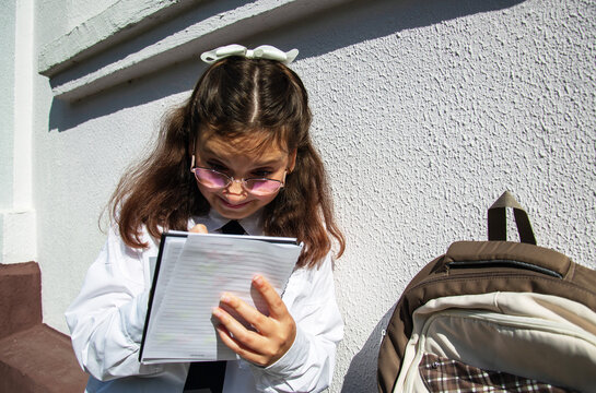 Schoolgirl sits outside her school and writes notes in a notebook while wearing glasses. She has a backpack beside her