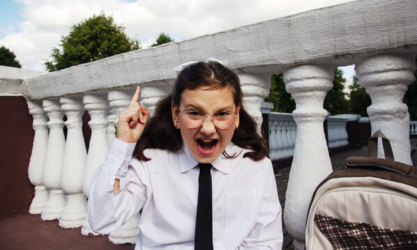 Student with glasses is raising one finger while making an expression of excitement outside a school. She wears a white shirt and a tie