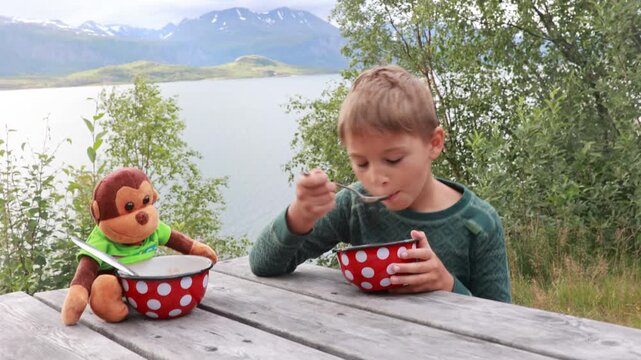 Children, boy brothers, having lunch in a reststop in Norway in beautiful nature. Kid with teddy bear eating lunch on a public wooden bench while on camping holiday in Norway, Lofoten