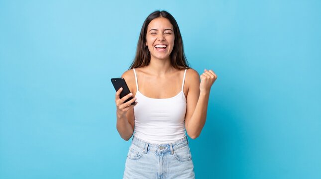 Happy young woman celebrating a win while looking at her smartphone isolated on a blue background