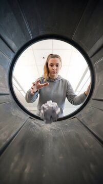 A young woman wearing a grey hoodie throws a crumpled piece of paper directly into a trash can. The shot is captured from an unusual POV inside the bin, highlighting themes of waste, failure, start...
