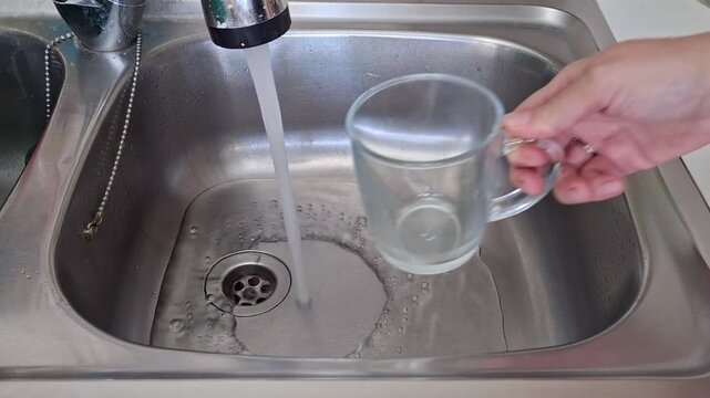 Water comes out from the tap and fills a cup in a kitchen sink while draining into the basin