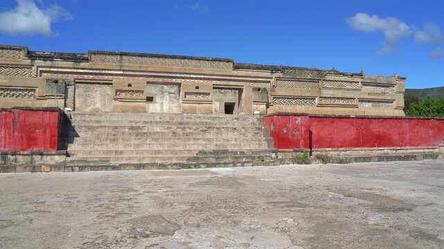 Pan wide shot of the main Zapotec palace facade with stone stairs and red stucco walls at Mitla archaeological site in Oaxaca, Mexico.