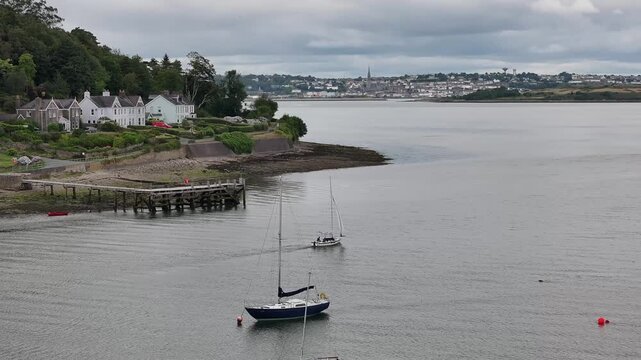 A calm aerial perspective of sailboats anchored in the harbor at Crosshaven, County Cork, featuring coastal houses and a wind turbine under an overcast sky.