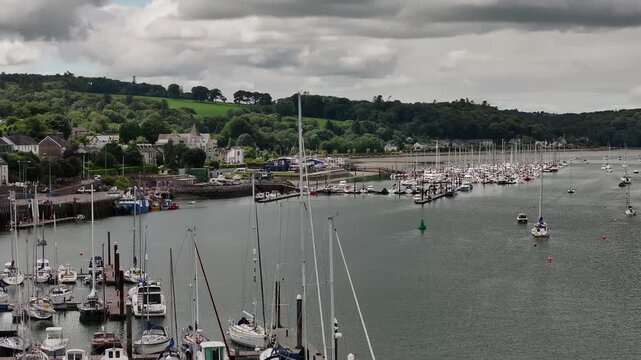A highangle drone shot capturing the serene harbor of Crosshaven in County Cork, Ireland, featuring numerous sailboats moored against a backdrop of lush green hills and a cloudy sky.