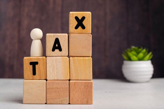 Wooden block tower spelling TAX diagonally with a small wooden peg figure standing on top, financial growth concept on dark background.