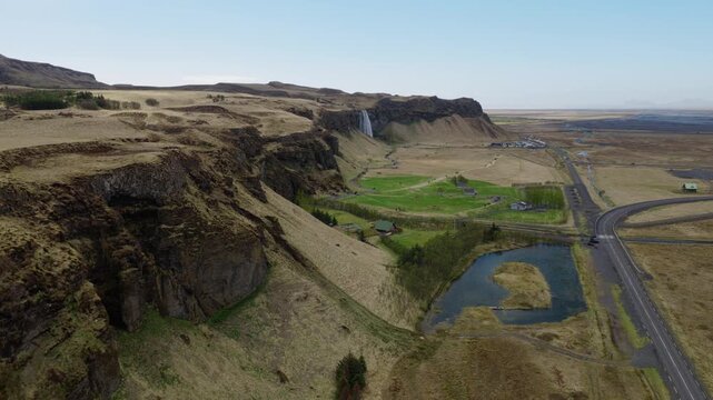 Seljalandsfoss area shows a basalt escarpment, Route 1, ponds and green farmland across the valley in South Iceland, drone flyover.