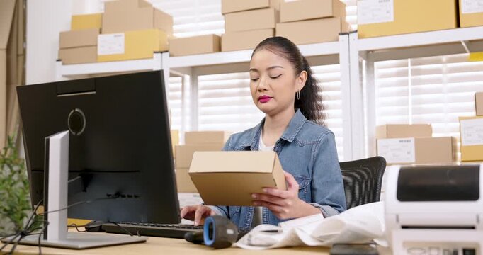 Asian woman small business owner lifting parcel box while reviewing order details, checking inventory list, managing e-commerce packaging process, preparing shipment workflow for online store delivery
