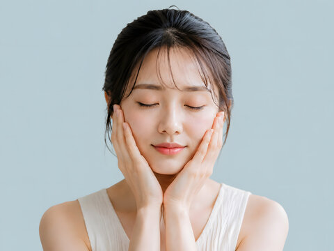 両手で頬に触れながら目を閉じるタンクトップを着た美肌の若い日本人女性