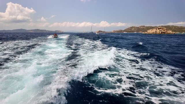 Panoramic view from the stern of a motorboat sailing through a stormy, dark blue sea, leaving behind a wide, dynamic trail of white, foaming water, with a mountainous coastal landscape