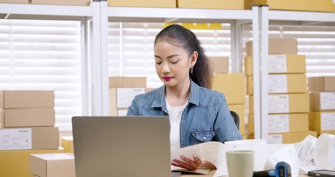 Asian woman small business owner scans printed order list beside laptop while reviewing e-commerce documents and organizing parcel paperwork for customer delivery