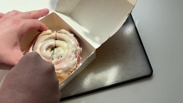 Hands slicing sweet cinnamon roll bun with cream glaze using knife in white takeaway box for food delivery concept