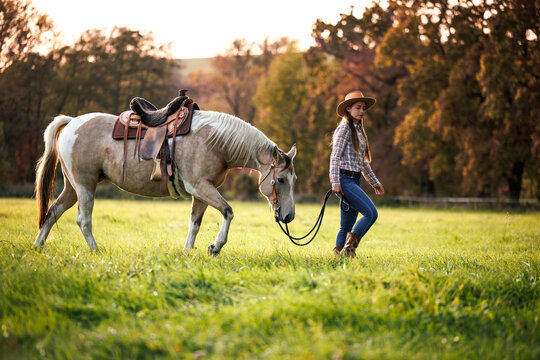 Cowgirl leading horse across pasture in autumn farm. Woman walking with horse, symbolizing slow living and country lifestyle