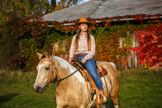 Cowgirl riding horse near stable covered red ivy creeper leaves at ranch in autumn. Woman wearing western clothing and cowboy hat