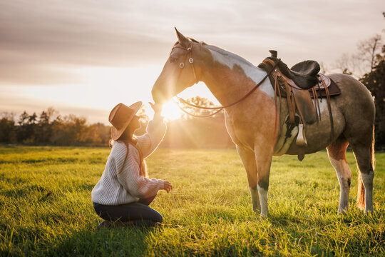 Relaxed woman resting beside her horse after horseback riding on pasture during sunset. Cowgirl enjoying western lifestyle at autumn countryside