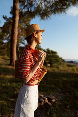Naklejka premium Young woman enjoying lifestyle at sunset in nature. She wears a straw hat and striped shirt. Concept of summer vacation, travel, relaxation, leisure, and outdoor hobby.